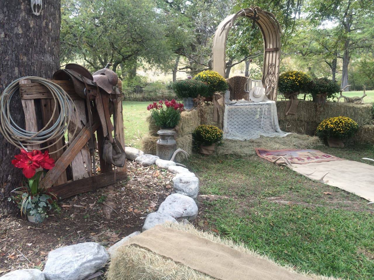 Rustic wedding ceremony arbor with burlap draping, antlers, hay bales, and mums leading to a lace-covered table at Sabinal River Lodge