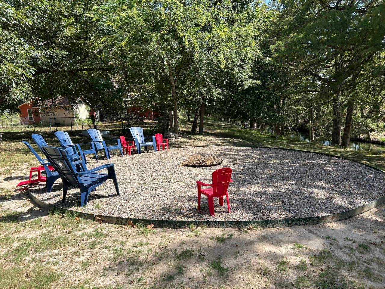 Riverside outdoor gathering area at Sabinal River Lodge with blue and red Adirondack chairs circling a fire pit near the creek in Utopia, Texas
